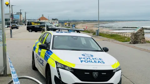 Police tape across the beach-side path in Aberdeen. Two police cars are visible, as well as uniformed officers.