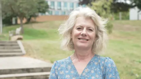 A woman standing outside. She has light hair and is wearing a blue top with flowers on it. Behind her is a grass bank and a stone staircase.