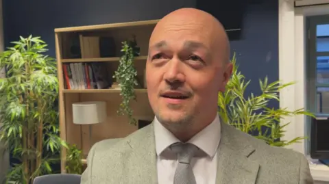 Graeme Littlejohn, a bald man with stubble, is wearing a light suit jacket with a white shirt and grey tie. He is in an office space with plants, a bookcase and a window behind him.