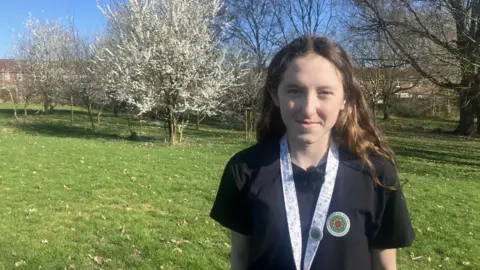 Young girl with long brown hair stands in the park wearing a black polo shirt.