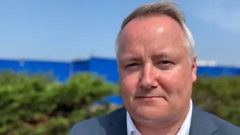 Darren Millar looks at the camera. He is clean shaven, with short grey hair, and is wearing a grey suit and white shirt. A hedgerow can be seen behind him. It is a head and shoulders shot of him.