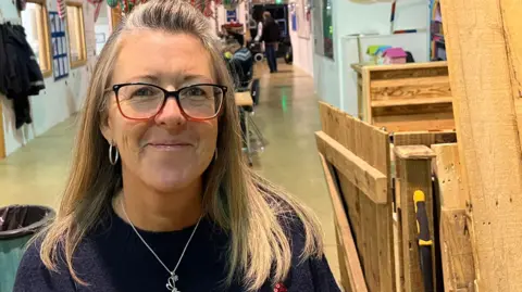 Sarah Lewis stands in the workshop next to wooden gates. She has blonde, shoulder-length hair and is wearing a Christmas jumper and glasses. Behind her is a long table with people sitting down and inflatable Christmas decorations overhead.