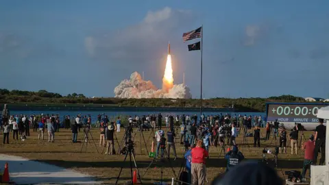 Chip Somodevilla/Getty Images Artemis is taking off in the distant background, and people watch, many with tripods. The US flag flutters above them on a flag pole.