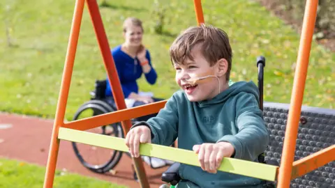 Fleur Bryant A child in a wheelchair wearing a hoodie. He is sat in a adapted swing. A woman in a wheelchair is behind him, looking and smiling.