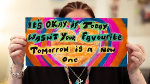 Supplied Woman holding colourful artwork portaying the message "It's okay if today wasn't your favourite, Tomorrow is a new one"