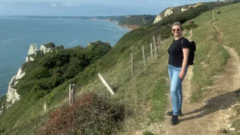 Lizzy Willmington smiling at the camera while walking on a coastal path overlooking the sea