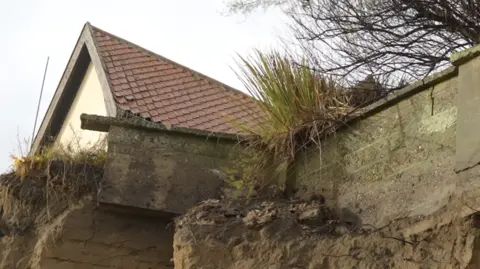 Jamie Niblock/BBC A house's foundations can be seen out the edge of a cliff that has eroded away. The home's roof can be seen just above the cliff on a grey day.