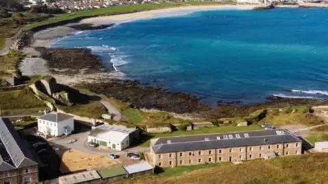 A row of buildings to the right can be seen with cliffs in the background and further detached buildings to the left with the sea stretching out behind. 