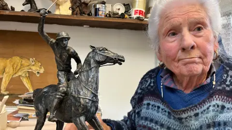 A brass sculpture of a polo player on a running horse sits next to Jan, who is in her 80s, in her studio at home in Stawell. 