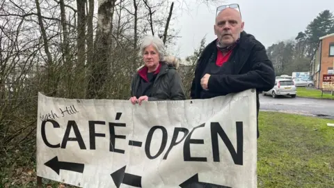 Debbie and John photographed with the sign for their cafe. Debbie is wearing a grey winter coat with a dark red and black fleece, and John is wearing a black coat with a black apron underneath. The flexible banner is on a grassy verge, and is white with black writing, reading "Heath Hill Cafe - Open" in large letters with three black arrows underneath. 