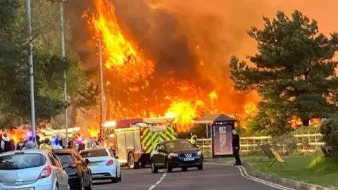 Nicola Lockyer A fire can be seen in the distance, with fire engines, cars and people in the foreground