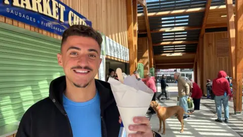 Andrew Turner/BBC David Cordoso has dark medium length hair and defined eyebrows, and a small beard and moustache. He is wearing a black jacket and a blue T-shirt underneath. He has a cone of chips in his hand, standing near the chip stalls in Great Yarmouth Market where there is a queue of people and a dog waiting in line at a stall.