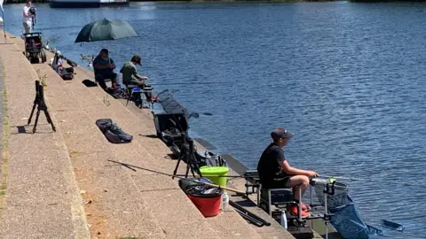 Three anglers, surrounded by equipment, sit on a series of concrete steps by a river