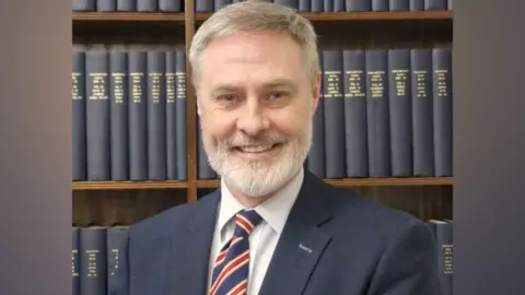 Paul Frew A man with white hair and beard, he has a navy suit, white shirt and red white and blue tie