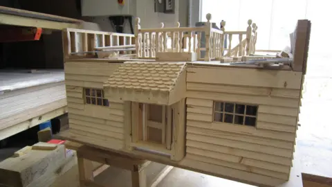 Alex Pope/BBC A large wooden toy house, with a door, windows and roof. Shelves are to the right and a doorway to the left. 