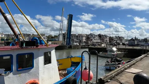 Maryport's Elizabeth Dock, A shipping trawler is in the foreground on the right-hand side with two smaller vessels moored nearby. Beyond them, the Ellenfoot pedestrian bridge - a white-and-blue structure - is raised, pointing high into the air.