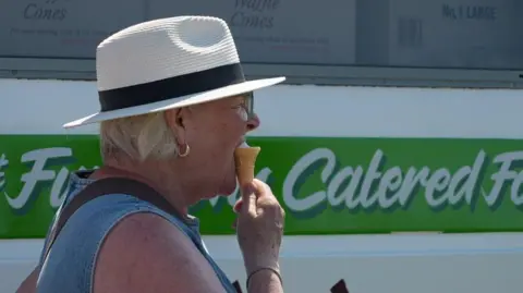A woman with blonde hair standing next to an ice cream van. She is wearing a straw hat and a denim top. She is turned to the side facing towards the right and is eating an ice cream in a cone.