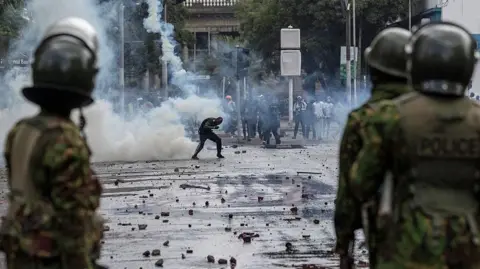 Police officers in camouflage uniform (foreground) and in the background a protester ducks a teargas canister in downtown Nairobi
