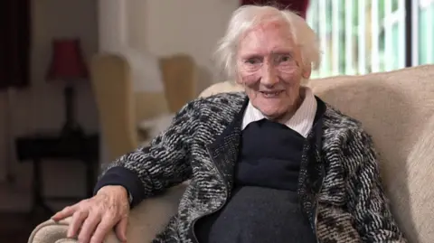 A woman with short white hair looks at the camera. She has a black and white knitted cardigan and has a black smock over a white collared shirt - the collars can be seen. She is sat on a cream sofa. She is in focus while the background is blurred.