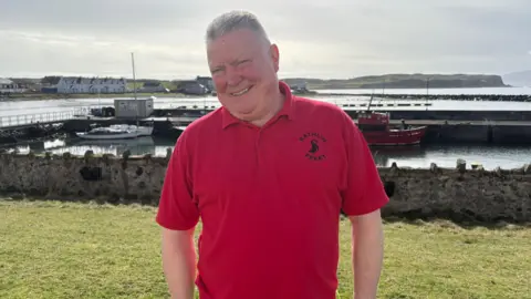 Tom is wearing a red t shirt that says Rathlin Ferry in black lettering. He's standing on grass, with the ocean behind him. Some boats are parked nearby and houses are across the port. 