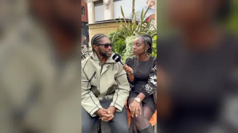 A man sat down outside by a plant with a woman next to him who is holding a mic to his mouth.