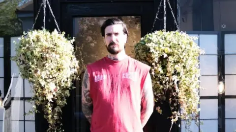 Adam Willett Adam Willett is wearing a red vest outside a shop, which has large hanging baskets of flowers by the door. He has brown hair, tattoos and a beard.