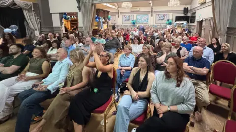 Crowds of people inside a function room sitting on chairs all facing forward.