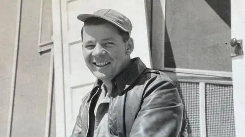 Bridgette Daily Joseph Gray wearing a leather jacket and soft cap on an angle, smiling. It is in black and white.