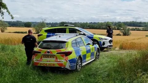 David Jackman/Everything Epping Forest Two police cars parked by the side of a large field. Two officers are looking into the field beyond them.