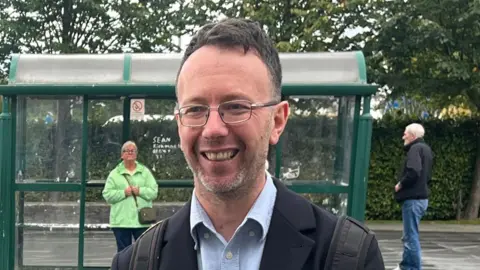 John Glenday has short dark hair and wears glasses while smiling off to the side. He stands in front of a road with a bus stop in the background