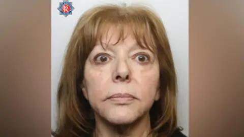 A woman with shoulder-length dark blonde hair is looking at the camera in a police mugshot. There is a Greater Manchester Police logo in the top left hand corner.