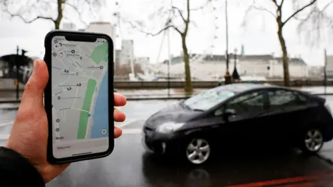 A person holding a phone with the Uber app open showing a map of London, with a black car on the road in the background