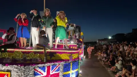 BBC A Jersey Battle of Flowers float drives towards the camera on the left of the image while crowds line the side of the street on the right. There are several people stood on the float which is decorated in multicoloured flowers. Four of the people appear to be dressed as the 1981 Eurovision winning band Bucks Fizz and are dancing.