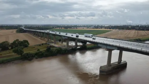 A landscape aerial photo of Ouse Bridge, which has several vehicles driving across it over water and fields beyond it.