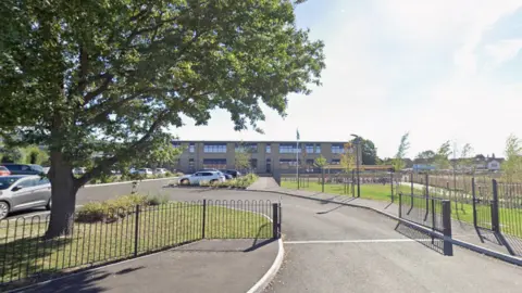 A school is seen from the gates. There is a big green tree next to the gate and the sky is blue.
