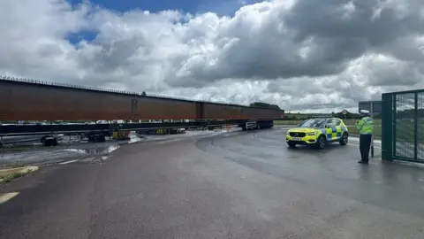 Northamptonshire Police A very long, large, metal beam, with a police car and a policeman to the right. There is a large gate. There is a large grey sky and a paved area.