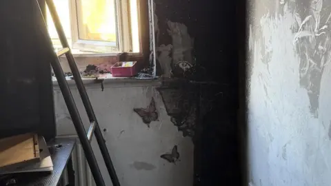 Essex County Fire and Rescue Service A blackened wall with butterfly decorations. There is smoke damage across the white wall and windowsill. A bed with desk underneath and ladder is visible.