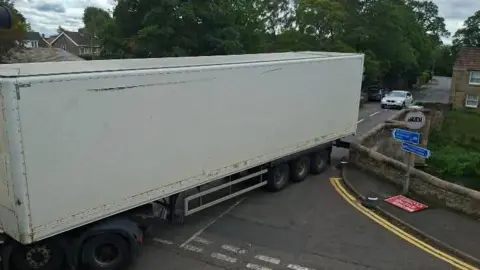 Paul Frith A large lorry with a white trailer squeezes across a narrow stone bridge, with trees and houses in the background. A sign advertises a 7.5-tonne weight limit