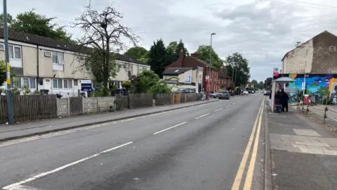 A street on an overcast day. There is a bus stop. There are terraced houses on the left side.