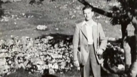 Family photo Gordon, aged 15, in slacks and a light jacket with a shirt underneath. He is outside standing on a wall next to a tree.