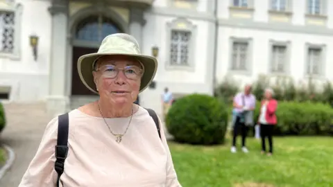 Angela is wearing a light pink blouse, green sun hat, and glasses stands in front of a large white building with multiple windows and an arched entrance. In the background, two people stand on the grass near bushes, and another person walks along a pathway.