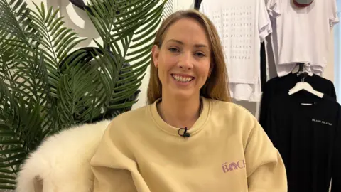 Catrin hughes sits on a white fur chair, wearing a yellow sweatshirt. She has light blonde/ brown hair.
