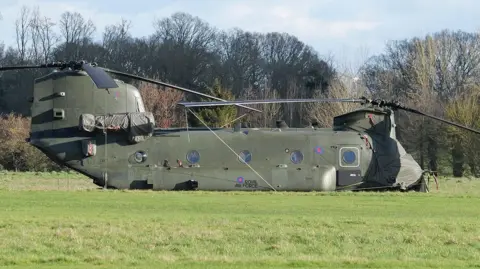 Richard Thomas A large khaki green Chinook landed on a field in front of trees. It has RAF branding on its side and big, black rotor blades.