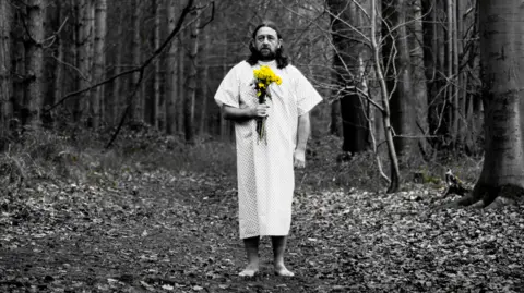 GenFest 2025 Lou Chawner is stood in the middle of the image in a forest. He is wearing a long medical gown and is holding a bunch of yellow flowers in his right hand. He has shoulder length hair and is looking towards the camera. 