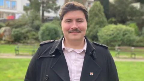 A man smiling and looking at the camera. He has brown hair and a mustache and is wearing a Guernsey pin. In the background, there are bushes and other greenery. 