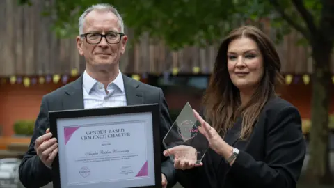 Prof Roderick Watkins and Fiona Drouet are standing next to each other. Watkins, who is on the left, has short grey hair and is wearing a blue shirt and a grey blazer. He is holding a certificate which is framed. Drouet, who is on the right, has long brown hair and is wearing a black blazer. She is holding a glass diamond shaped award.