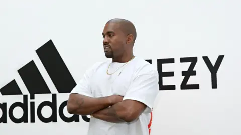 Getty Images West in a white T-shirt with arms folded in front of Adidas/Yeezy sign