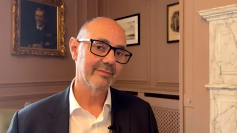 A bald man wearing black glasses and a suit looking into the camera. He is stood in a room with light pink walls and pictures hung behind him. To the right of the picture is the edge of a white fireplace
