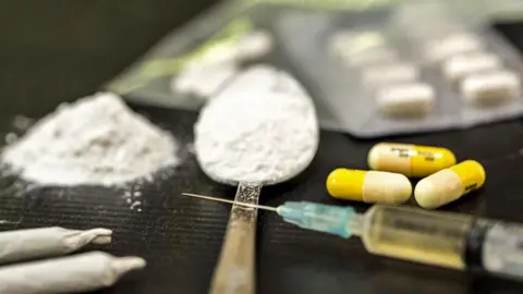 A table with drugs on including a spoon with white powder, a needle, pills and joints. 