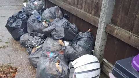 South Kesteven District Council About a dozen black bin bags, containing waste, left on concrete next to a brown, weathered wooden fence. An old white lampshade can also be seen.